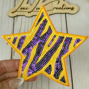 May include: A yellow star-shaped sequin patch with purple and black stripes. The patch has an orange border and is held by a hand. The wooden sign in the background says "Lexi Ju Creations".