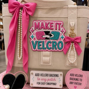 May include: A beige tote bag featuring a large embroidered patch that reads MAKE IT VELCRO in pink and teal. The bag is decorated with two bright pink satin bows and strings of white faux pearls. Informational signs about Velcro backing are visible.