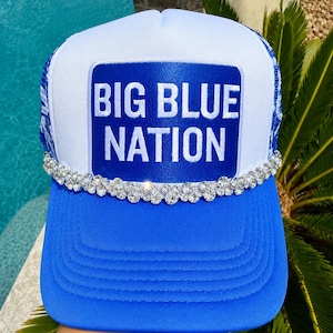 May include: A white and blue trucker hat featuring a rectangular blue patch with the white text BIG BLUE NATION. The front panel is accented with a horizontal row of clear, faceted rhinestones along the base of the crown above the blue brim.