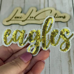 May include: A sequined patch in the shape of the word "eagles" in gold sequins, outlined with silver glitter. The patch is held in a hand, with a wooden sign in the background.