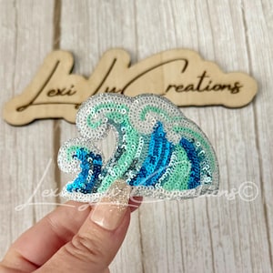 May include: A wave-shaped patch covered in shimmering sequins in shades of white, light green, teal, and dark blue. The design features a stylized cresting wave pattern with a distinct curved shape, crafted with dense, reflective sequin embroidery.