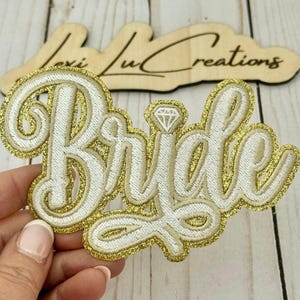 May include: White embroidered "Bride" patch with a gold glitter border and a diamond detail. The patch is in a cursive font. A wooden sign with the text "Lexi Lu Creations" is in the background.