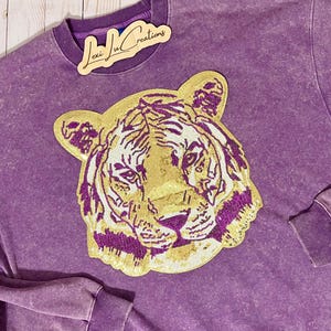May include: Purple sweatshirt with a gold and purple tiger applique. The sweatshirt has a distressed look and the applique is sparkly.