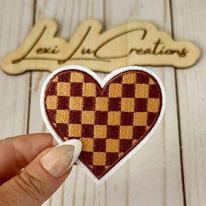 Op de afbeelding: Geborduurde hartvormige patch met een dambordpatroon in bruin- en goudtinten, omlijnd met een witte rand. De patch wordt vastgehouden voor een houten bord met de tekst "Lexi Ju Creations".