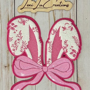 May include: Pink felt bunny ears with a large pink sequined bow. The ears have a white felt background with a pink floral pattern.