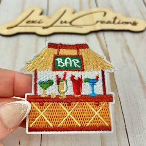 May include: Embroidered patch featuring a tropical bar scene. The patch depicts a thatched-roof bar with the word "BAR" on a green sign. The bar is decorated with colorful cocktails. The patch is held by a hand.
