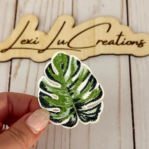 以下が含まれることがあります： 緑と白のモンステラ葉のデザインが施された刺繍パッチ。パッチは白い縁取りがあり、手で持たれています。背景の木製の看板には「Lexi Lu Creations」と書かれています。