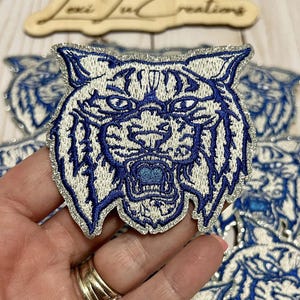 May include: A white and blue embroidered patch of a snarling wildcat with a silver glitter background.