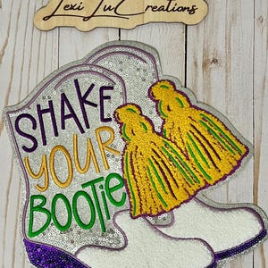 Może przedstawiać: Ozdobny znak z parą białych kowbojskich butów z napisem "SHAKE YOUR BOOTIE" w kolorze fioletowym, złotym i zielonym. Buty są ozdobione cekinami i żółto-zielonymi frędzlami. U góry drewniany znak z napisem "Lexi Ju Creations".