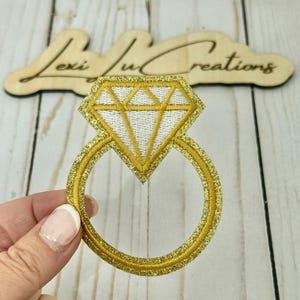 May include: A gold glitter embroidered ring patch with a white diamond-shaped center. The ring is held by a hand. The wooden sign in the background reads "Lexi Ju Creations".