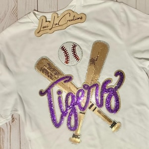 May include: White t-shirt with a baseball theme. Features a baseball, two crossed bats, and the word "Tigers" in purple sequins. The bats are embellished with gold sequins. A wooden sign with the text "Lexi Lu Creations" is at the top.
