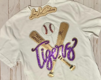 May include: White t-shirt with a baseball theme. Features a baseball, two crossed bats, and the word "Tigers" in purple sequins. The bats are embellished with gold sequins. A wooden sign with the text "Lexi Lu Creations" is at the top.