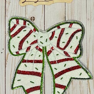 May include: A decorative Christmas bow with a white base, red stripes, and green and silver sequin trim. The bow has a textured appearance and is embellished with small green beads. The wooden sign above the bow reads "Lexi Lu Creations."