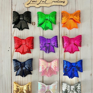 May include: Twelve sequined bows in various colors, including black, green, orange, red, purple, pink, blue, gold, white, and silver. The bows are arranged in three rows of four on a light wood background. The text "Lexi Lu Creations" is visible on a wooden sign above the bows.