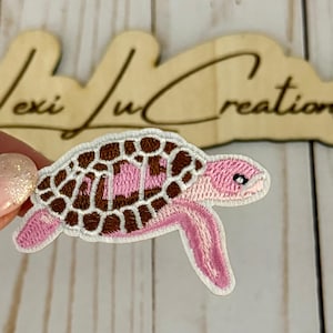 May include: Embroidered patch in the shape of a sea turtle. The turtle has a brown and pink shell, pink flippers, and a white border. The patch is held by a person's hand. The background has the text "Lexi Lu Creations".