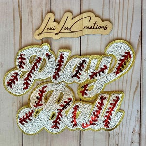 May include: Baseball-themed sequined applique with the words "Play Ball" in a script font. The letters are white with red stitching and gold glitter edging. A wooden sign with the text "Lexi Ju Creations" is above.