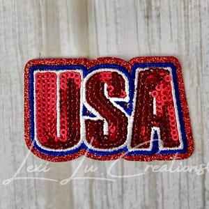 May include: A rectangular patch featuring the letters USA in red sequins with a white border and a blue outline. The entire patch is framed by a red sequined border, creating a bold, patriotic design with a textured, shimmering finish.