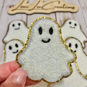 May include: White ghost-shaped felt applique with a gold glitter border and black facial features. The image shows several of these appliques, with one held in a hand. The text "Lexi Lu Creations" is visible in the background.