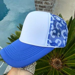 May include: A trucker hat featuring a solid white foam front panel, a bright blue curved brim, and a blue and white mesh back panel with a leopard print pattern. The hat has a structured crown and a white cord trim along the base of the front panel.