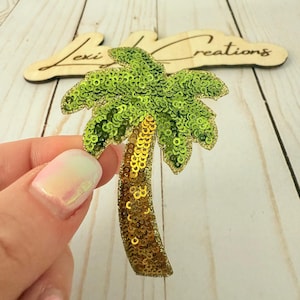 Peut inclure: Applique de palmier à paillettes avec un tronc doré et des frondes vertes. L'applique est tenue devant une enseigne en bois qui dit "Lexi Creations". Le palmier est une fourniture artisanale sur le thème tropical.