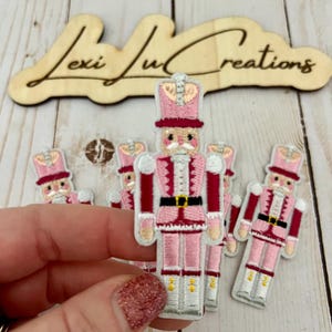May include: Five pink and red embroidered nutcracker ornaments with white accents. The nutcrackers are holding a drum and a stick.