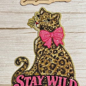 May include: A sequined leopard patch featuring a gold and black animal print, a bright pink bow at the neck, and the text STAY WILD in pink sequins at the bottom. Above the leopard is a wooden cutout sign with the text Lexi Lu Creations.