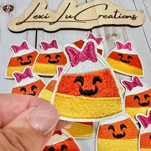 May include: Embroidered candy corn patches with smiling faces and pink bows. The patches feature white, orange, and yellow colors, perfect for Halloween crafts. A wooden sign with the text "Lexi Lu Creations" is in the background.