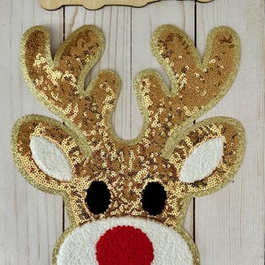 May include: A festive reindeer applique with gold sequins, white accents, a red nose, and black eyes. The antlers are covered in gold sequins. The wooden sign above reads "Lexi Ju Creations".