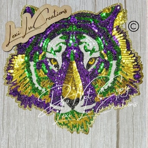 May include: A sequined tiger head in purple, green, and gold. The design features a detailed face with yellow eyes and a white muzzle. The image includes the text "Lexi Lu Creations" and "Do Not Copy".