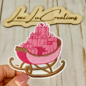 May include: Embroidered patch featuring a pink sleigh filled with stacked pink gift boxes tied with white bows. The sleigh has brown runners and a pink interior. The patch is on a white background. The text "Lexi Lu Creations" is above the patch.