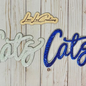 May include: Two iron-on patches with the word "Cats" in a cursive font. The left patch is white with silver sequins and the right patch is blue with silver sequins.
