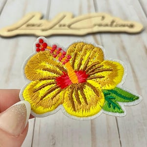 May include: Embroidered yellow hibiscus flower patch with red and orange accents. The patch has green leaves and a white border. The patch is held by a hand.