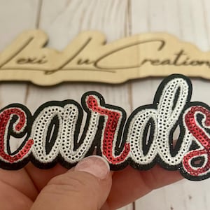 May include: A red and white sequined iron-on patch that says "cards" in a cursive font.