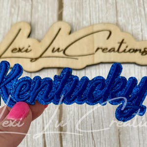 May include: A blue glittery "Kentucky" patch with a cursive font is held in front of a wooden cutout with the text "Lexi Lu Creations". The patch is a vibrant blue color and has a glittery texture.
