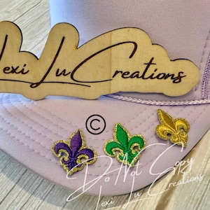 May include: A light purple trucker hat with a wooden Lexi Lu Creations logo and three fleur-de-lis patches in purple, green, and gold. The hat has a mesh back and a curved brim.