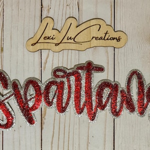 May include: Wooden sign with the text "Lexi Lu Creations" above a red and silver sequined "Spartans" sign. The signs are on a light wood background.
