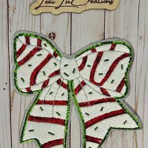 Pu&ograve; includere: Un grande fiocco decorativo con base bianca, strisce rosse e dettagli verdi. Il fiocco &egrave; bordato con paillettes verdi e ha un centro argentato. Il testo "Lexi Ju Creations" &egrave; visibile in alto.