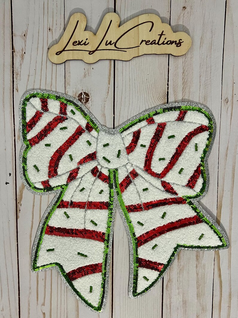 Pu&ograve; includere: Un grande fiocco decorativo con base bianca, strisce rosse e dettagli verdi. Il fiocco &egrave; bordato con paillettes verdi e ha un centro argentato. Il testo "Lexi Ju Creations" &egrave; visibile in alto.