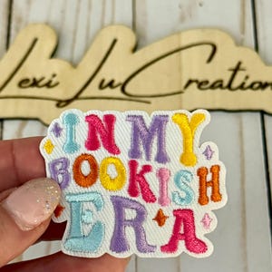 Puede incluir: Parche bordado con la frase "IN MY BOOKISH ERA" en letras coloridas. El parche tiene un fondo blanco, un borde azul claro y pequeños acentos de estrellas. Un letrero de madera con el texto "Lexi Lu Creations" está en el fondo.