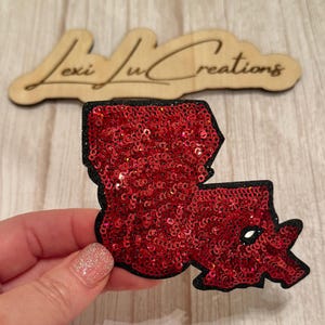 May include: A red sequined Louisiana state-shaped patch with a black border. The patch is held in front of a wooden sign that reads "Lexi Lu Creations". The sequins sparkle, reflecting light. The patch is likely for clothing or accessories.