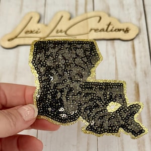 May include: A black sequined Louisiana state-shaped patch with a gold glitter border. The patch is held in a hand, with a wooden sign in the background that reads "Lexi Ju Creations".