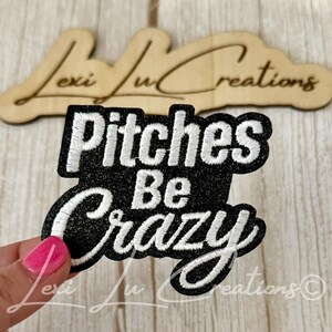 May include: Black embroidered patch with the words "Pitches Be Crazy" in white script. The patch is in a unique shape and is likely for clothing or accessories. A wooden sign with the name "Lexi Lu Creations" is in the background.