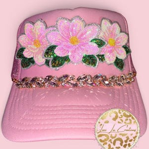 May include: Pink trucker hat with a floral design. The hat features three large pink sequined flowers with green sequined leaves and a sparkling crystal band. A circular logo with gold text and a leopard print is on the side.
