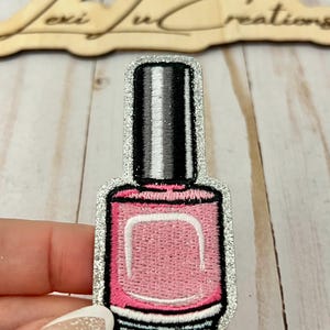 May include: A silver glitter iron-on patch of a pink nail polish bottle with a black cap.