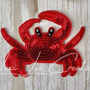 May include: A red sequined crab applique with black and white eyes. The crab has a detailed design with visible stitching and is set against a light-colored background. The applique is likely for clothing or craft projects.