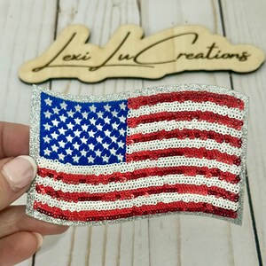 May include: A sequined American flag patch with a silver glitter border. The flag features red and white stripes and a blue field with white stars. The wooden sign in the background reads "Lexi Ju Creations".
