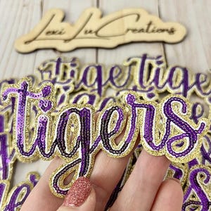 Op de afbeelding: Paarse en gouden "Tigers" woorduitsparingen met pailletten. De letters zijn omlijnd met gouden glitter en zwart, met een paarse paillettenvulling. Het woord is in cursief geschreven. Een houten bord met de tekst "Lexi Lu Creations" staat op de achtergrond.