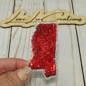 May include: A red sequined patch in the shape of the state of Mississippi with a silver glitter border. The patch is held in a hand, with a wooden sign that reads "Lexi Lu Creations" in the background.