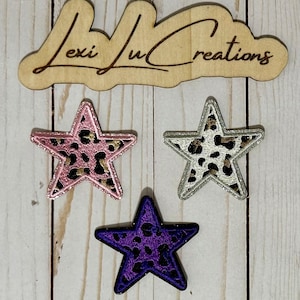 Pode incluir: Três patches em forma de estrela com centros de estampa de leopardo. Um é rosa com uma borda dourada brilhante, outro é prateado e o terceiro é roxo. Uma placa de madeira com o texto "Lexi Lu Creations" está acima dos patches.