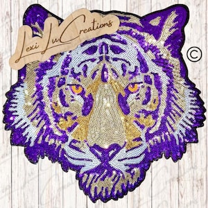 May include: A sequined tiger face in purple, gold, and white. The tiger's face is detailed with orange eyes and a neutral-colored snout. The design is embellished with sequins and has a wooden sign that says "Lexi Lu Creations".